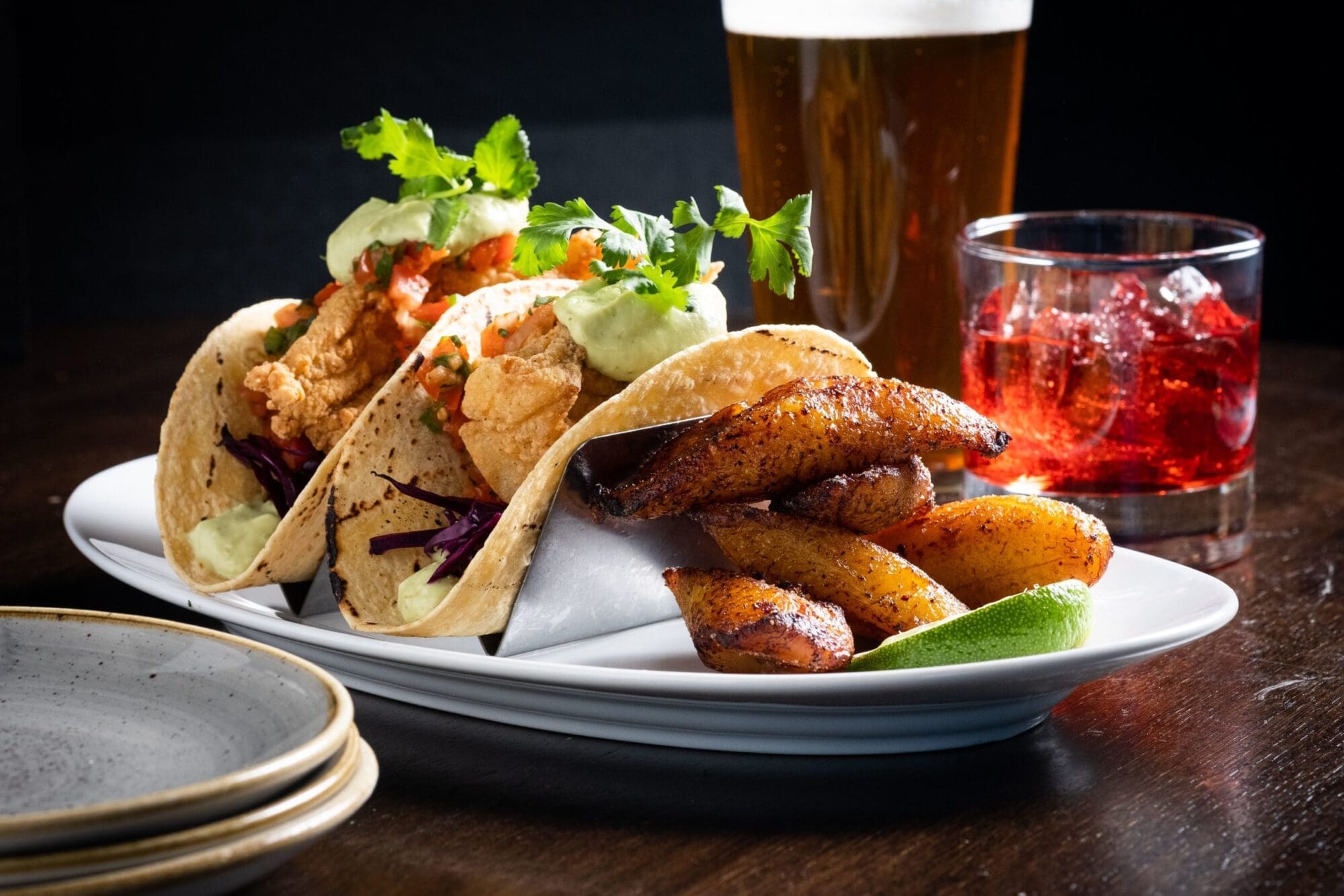 Fish tacos with lettuce, limes and pico de gallo with beer and cranberry juice in background.