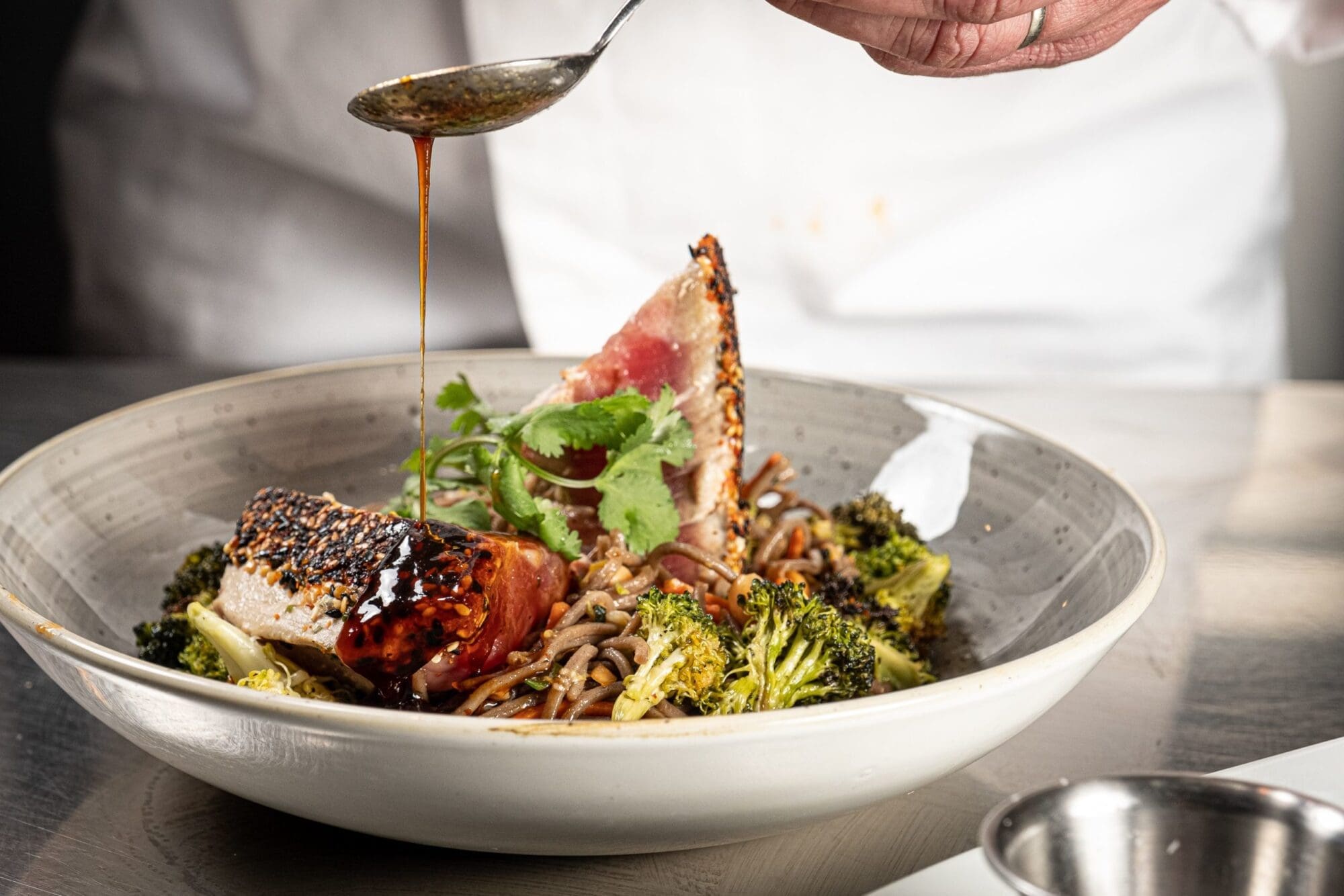 A chef drizzles sauce over a plated dish featuring seared tuna garnished with herbs, arranged on a bed of soba noodles and roasted broccoli. The dish is presented in a large, shallow bowl on a grey surface.