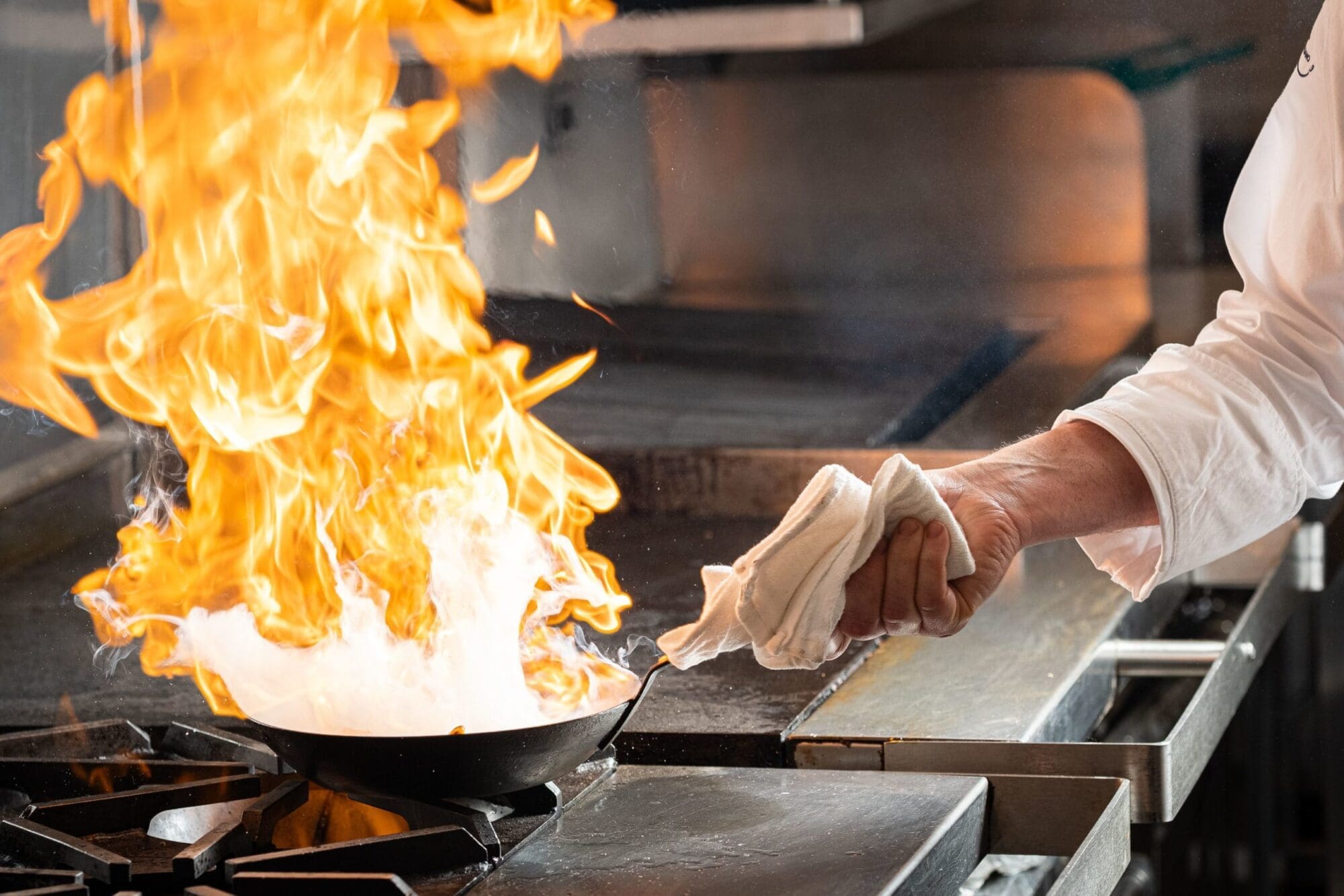 A chef's hand holding a towel and gripping the handle of a skillet with a large flame rising from it. The scene takes place in a commercial kitchen with a stovetop and other cooking equipment visible in the background.