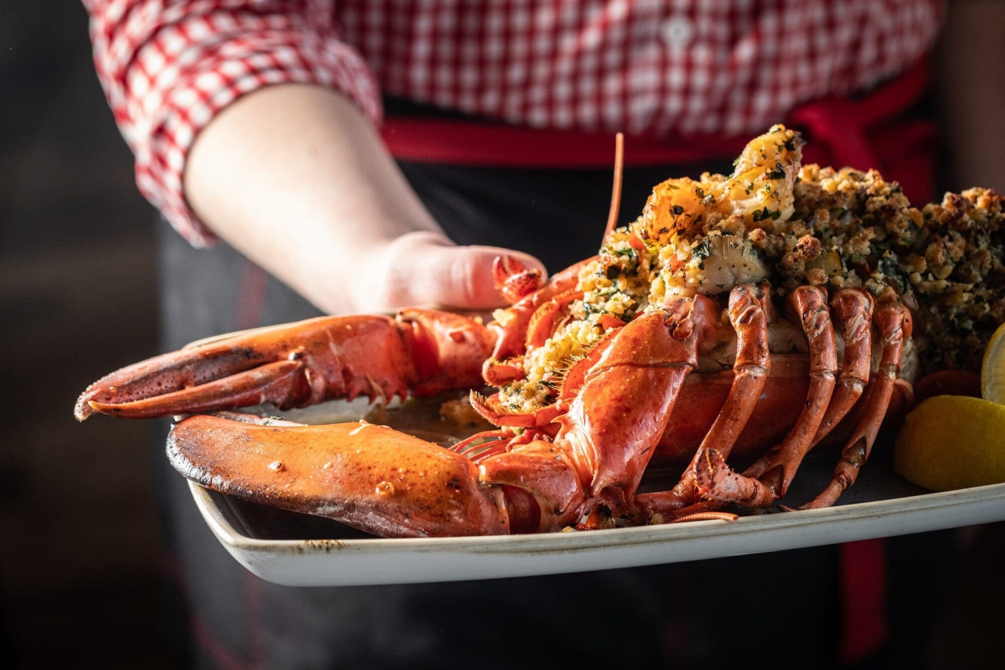 A person in a red and white checkered shirt and a red apron presents a dish featuring a large, garnished lobster on a white rectangular plate. The lobster is topped with a breadcrumb and herb mixture, and a lemon wedge is seen on the side.