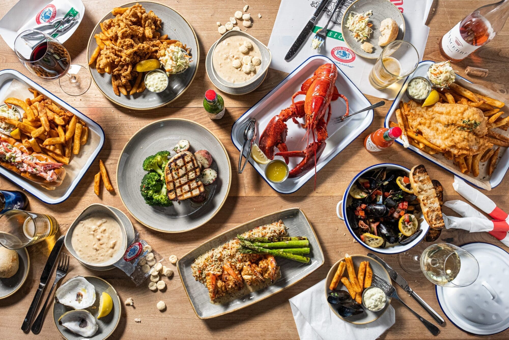 A wooden table covered with various seafood dishes, including a whole lobster, fried shrimp, mussels, grilled salmon, clam chowder, fish and chips, and side items like fries, coleslaw, and bread rolls. Beverages and condiments are also present.