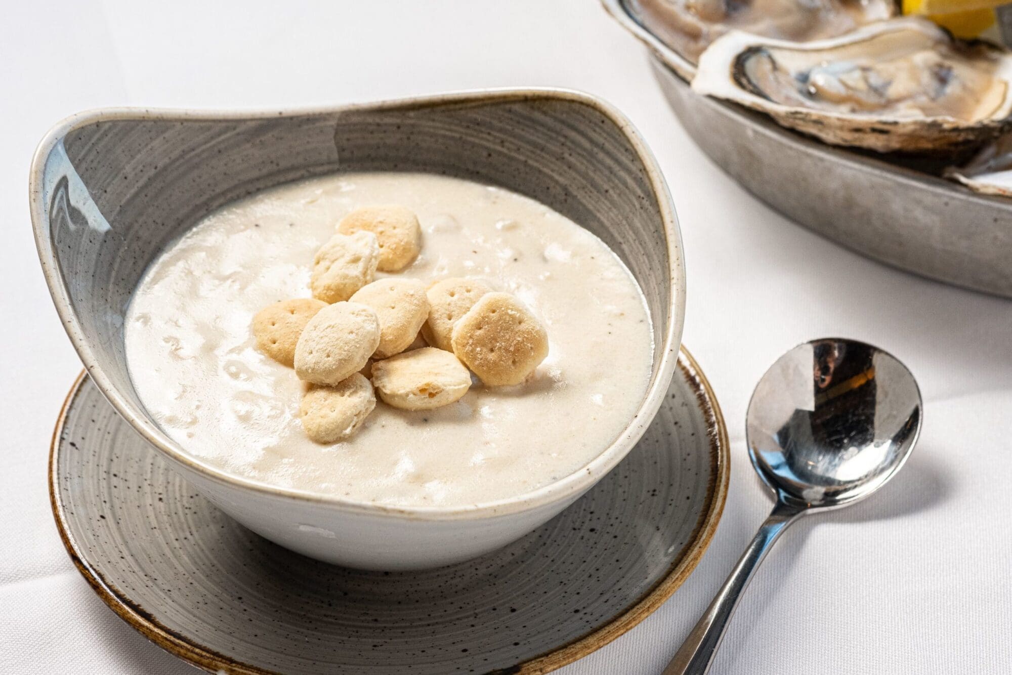 A bowl of creamy clam chowder topped with oyster crackers is served in a rustic dish on a saucer. A spoon is placed next to the bowl, and a plate with open oyster shells is partially visible in the background. The setup is on a white tablecloth.