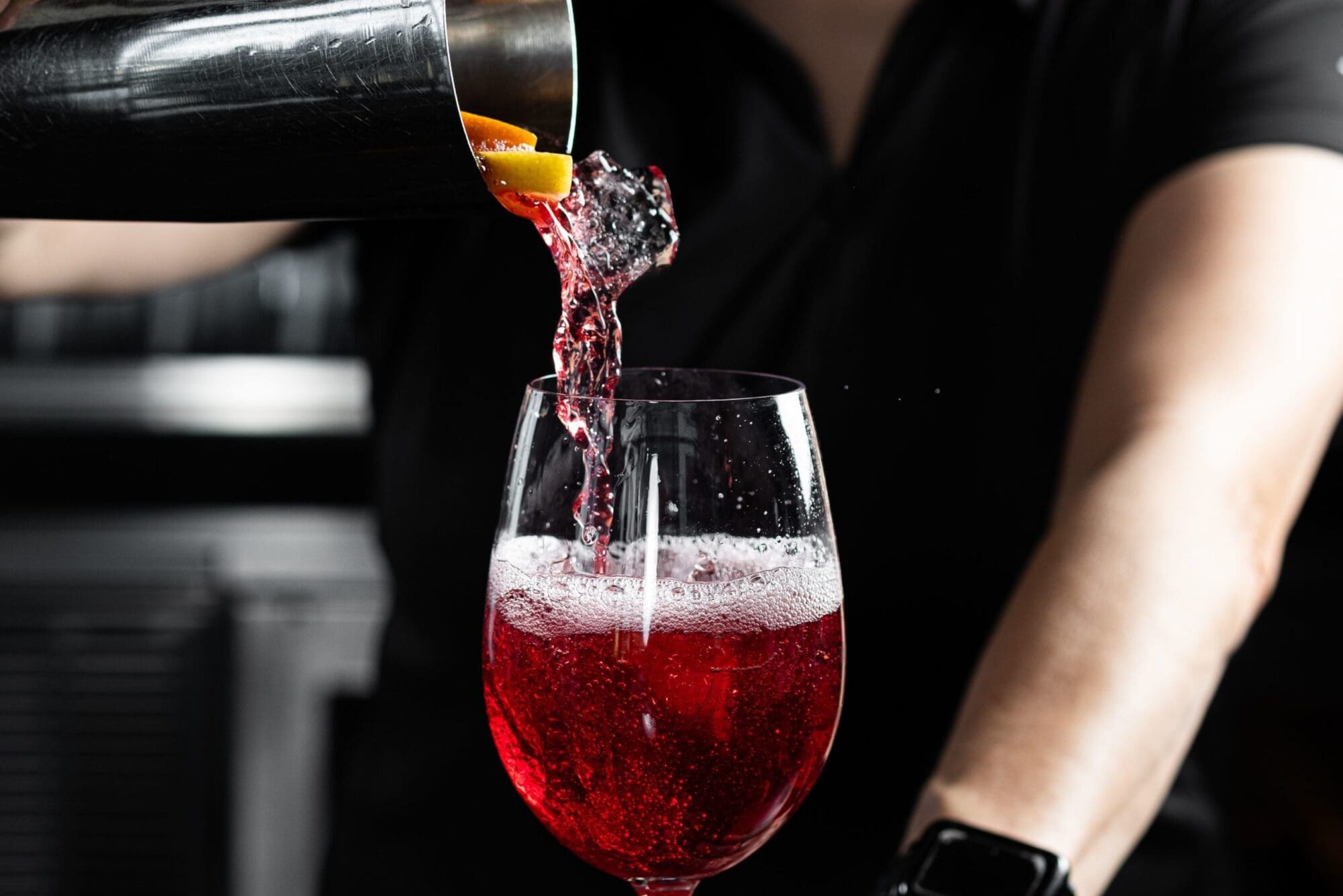 Bartender pouring a red cocktail from a shaker into a wine glass, with an orange slice visible in the mix. The background is out of focus, highlighting the drink preparation.