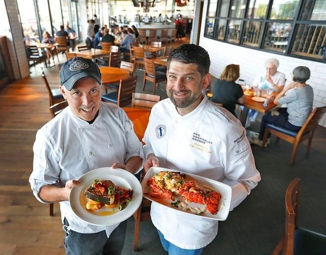 Two chefs proudly display their dishes inside a bright, busy restaurant. Both chefs wear white uniforms and smile at the camera. One holds a plate of seafood pasta, while the other presents a large lobster dish. Diners are seated at wooden tables in the background.