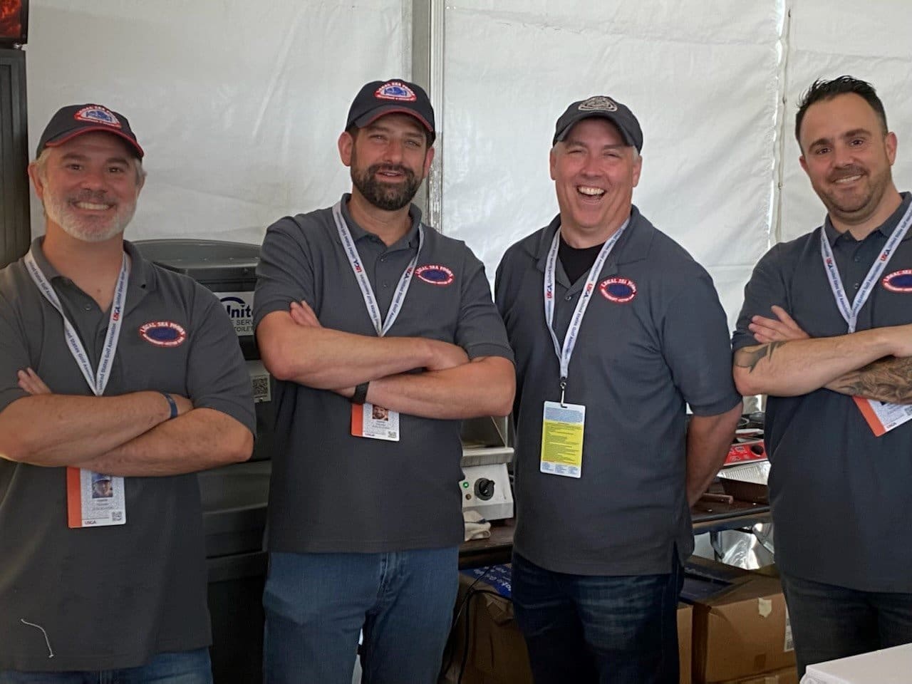 Four men are standing together, smiling and posing for the camera in a casual, relaxed manner. They are all wearing matching gray polo shirts with a logo, lanyards with ID badges, and caps. It appears to be in an event setting with equipment in the background.