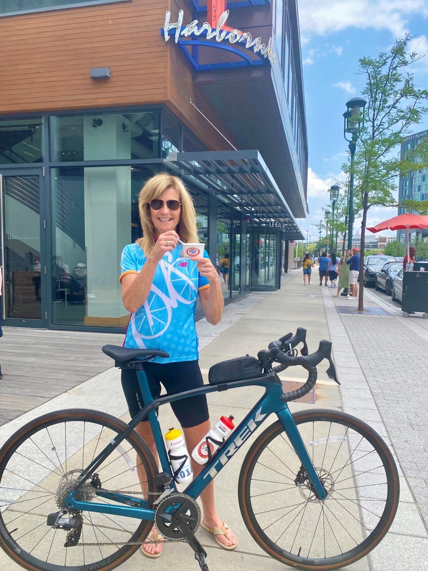 A woman in a bright blue cycling jersey stands next to a bike on an urban street. She is holding a cup with a smile. The bike is a Trek model with water bottles in the holders. Shops and buildings are in the background on a clear, sunny day.