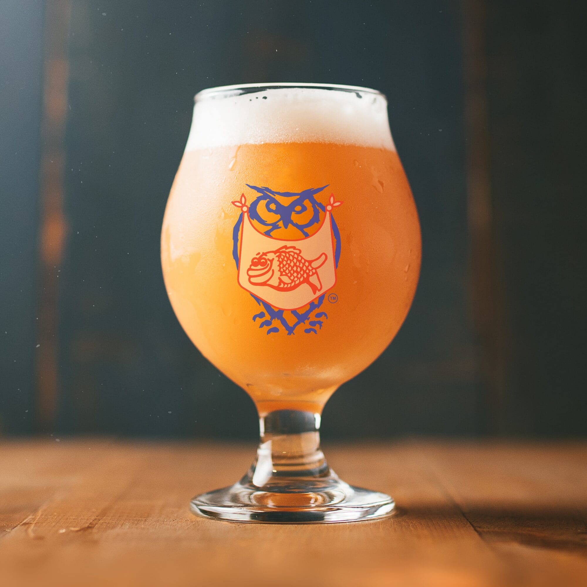 A tulip glass filled with a hazy, amber-colored beer sits on a wooden surface. The glass features a playful owl logo with outstretched wings, holding a fish in its talons. Light foam tops the beer, and the background is softly blurred.