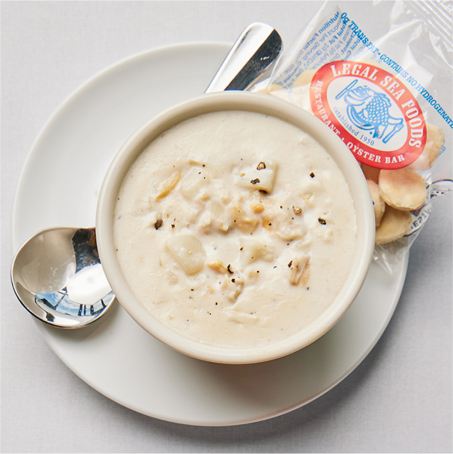 A white bowl filled with creamy clam chowder, sprinkled with black pepper, is placed on a white plate with a metal spoon. A packet of oyster crackers from Legal Sea Foods is next to the bowl on the plate. The background is a plain table surface.