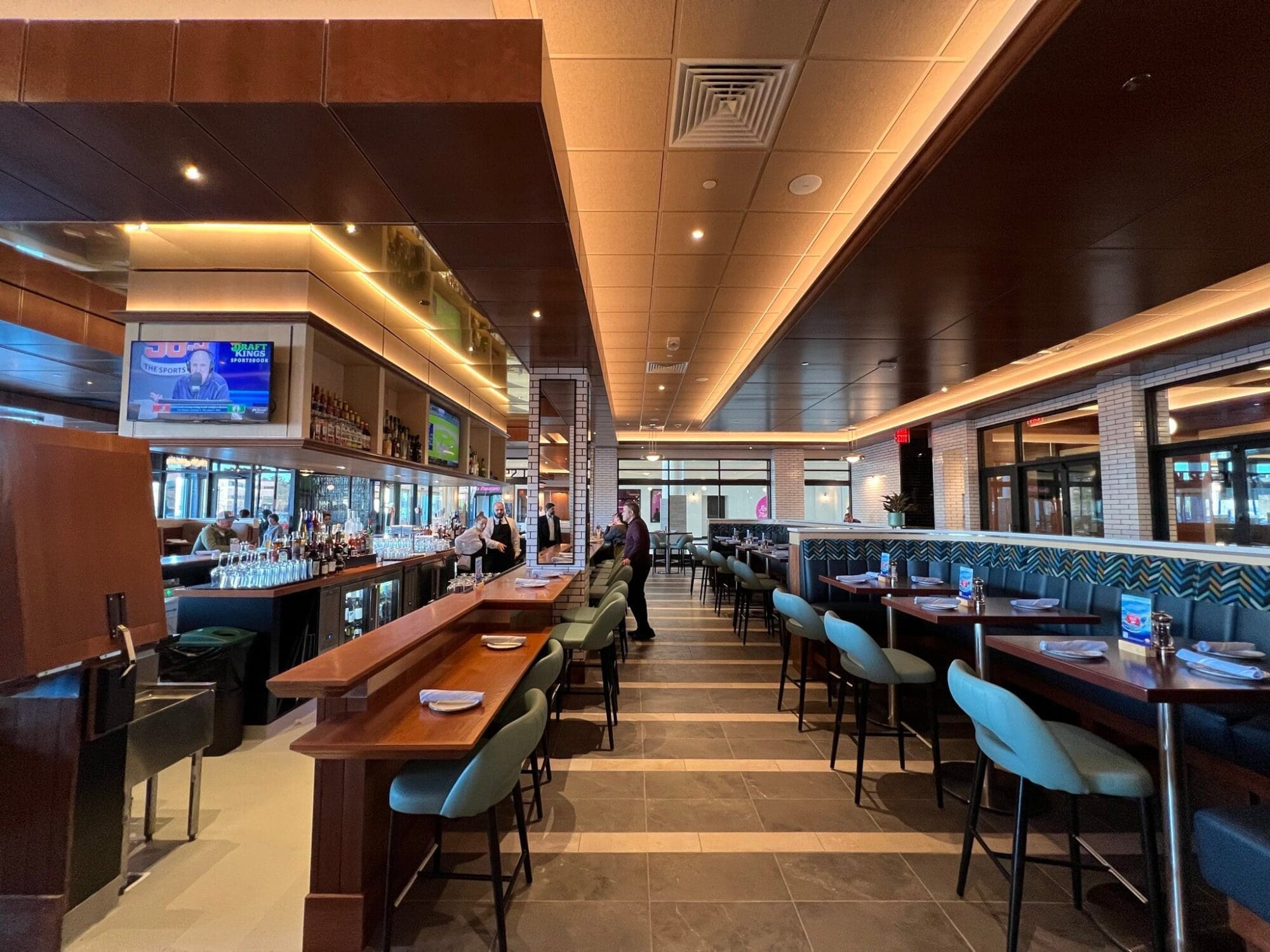 A modern restaurant interior with wooden tables, green chairs, and a bar area on the left. The ceiling has recessed lighting, and a TV screen is visible above the bar. Two employees stand behind the bar, and the dining area is empty.