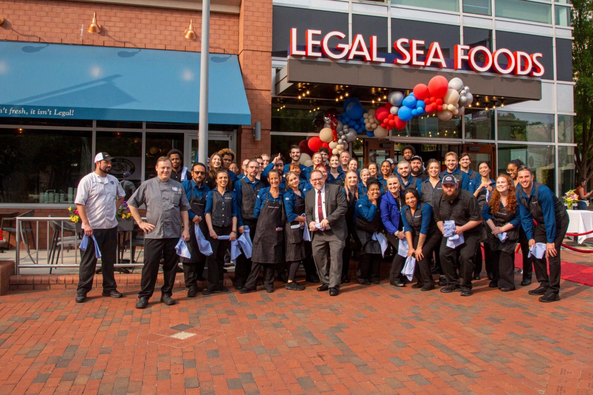 A large group of individuals, some in chef uniforms and others in formal attire, pose together in front of a restaurant named 