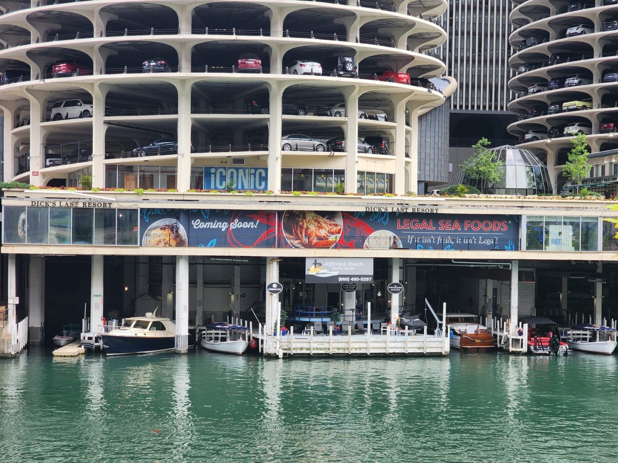 A waterfront scene showing a multi-level parking structure with vehicles parked on various levels. Boats are docked in the water below. Two prominent signs read 