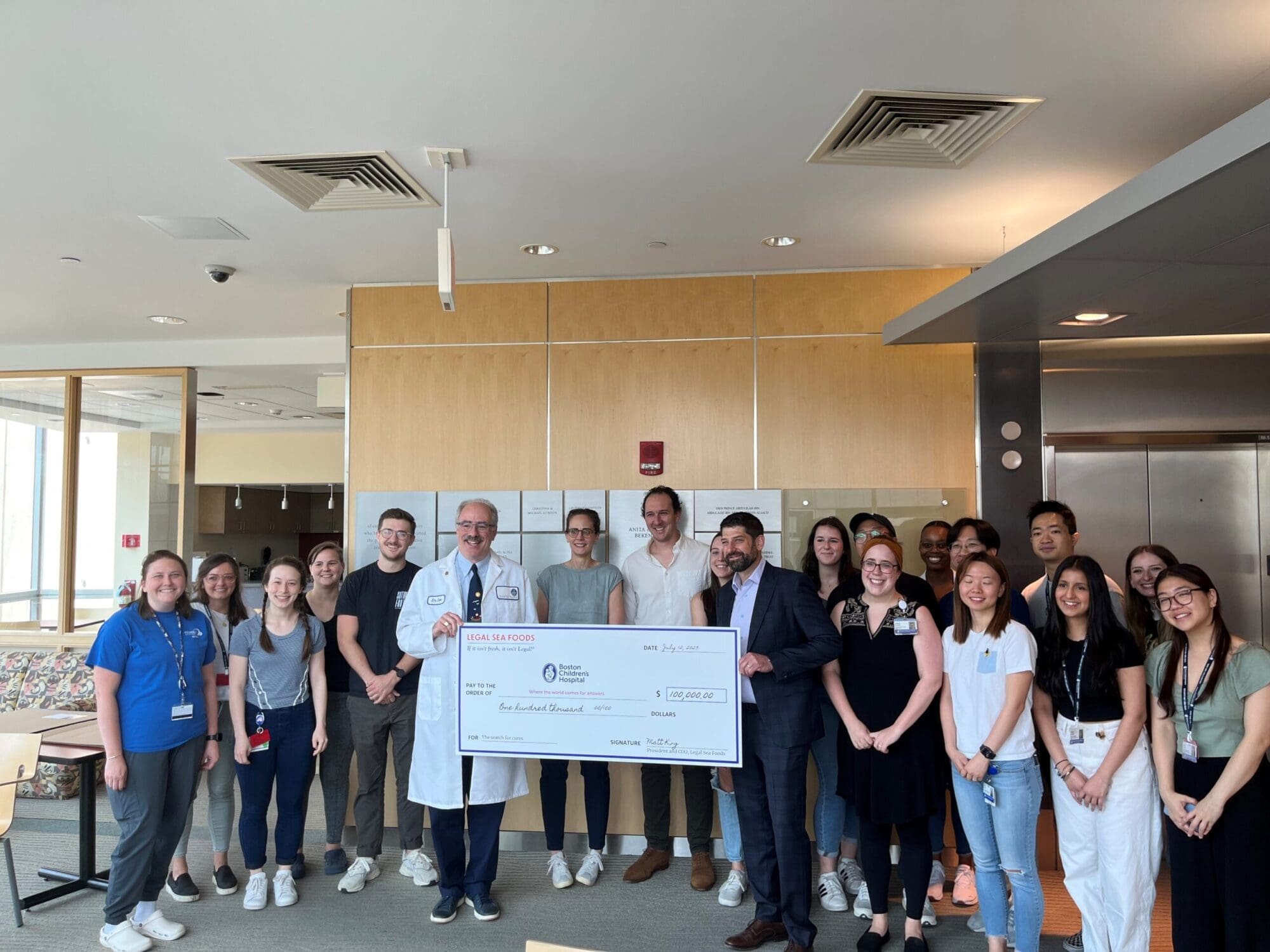 A group of people stand together indoors, smiling and holding a large ceremonial check. The group includes individuals dressed in casual clothing, business attire, and medical uniforms. They appear to be in a bright, modern room with wood paneling and large windows.