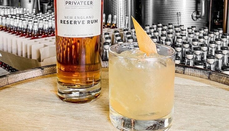 A bottle of Privateer New England Reserve Rum stands on a wooden surface in front of a background filled with rows of other bottles. Next to it, a glass filled with a light-colored cocktail garnished with a citrus twist rests on the same wooden surface.