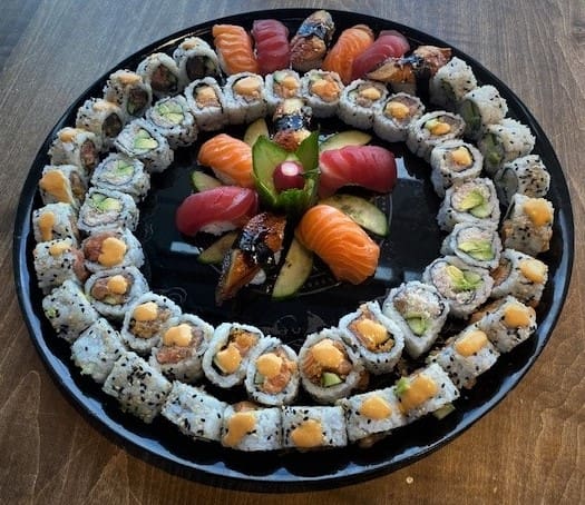 A round black platter filled with a variety of sushi. The outer ring consists of rolls topped with orange sauce, and the inner ring has different rolls. In the center are pieces of nigiri with toppings like salmon and tuna, garnished with cucumber slices.