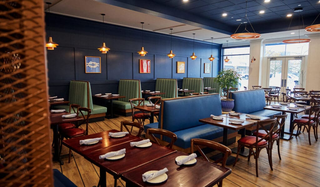 A modern restaurant interior with dark blue walls and wooden flooring. Green and blue booth seating line one wall, with wooden chairs and tables throughout. Pendant lights hang from the ceiling, and art pieces adorn the walls. Tables are set with white napkins.