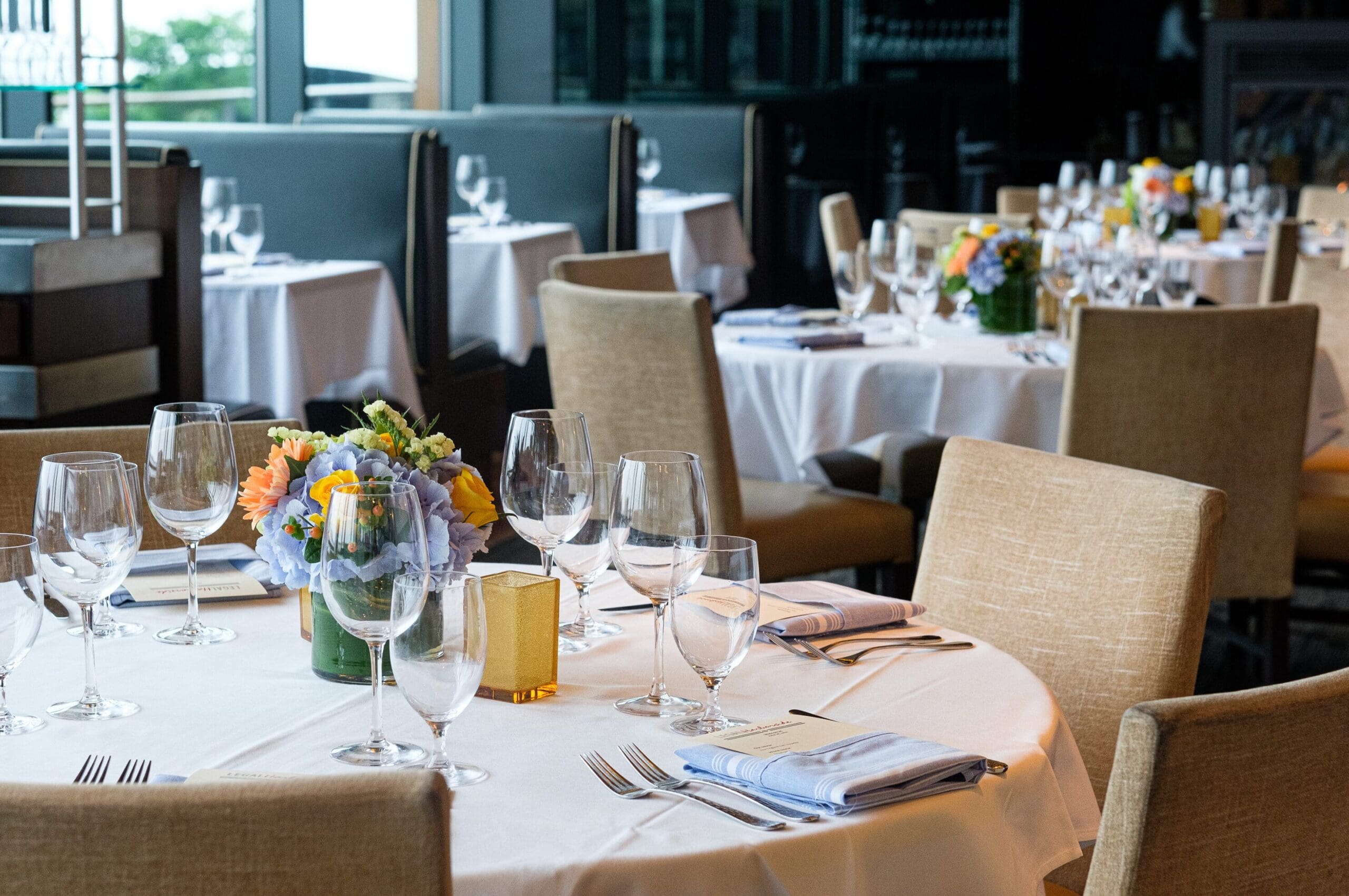 Elegant table settings with neatly arranged wine glasses, water glasses, silverware, and folded blue striped napkins on white tablecloths. Blurred background shows additional tables with similar arrangements