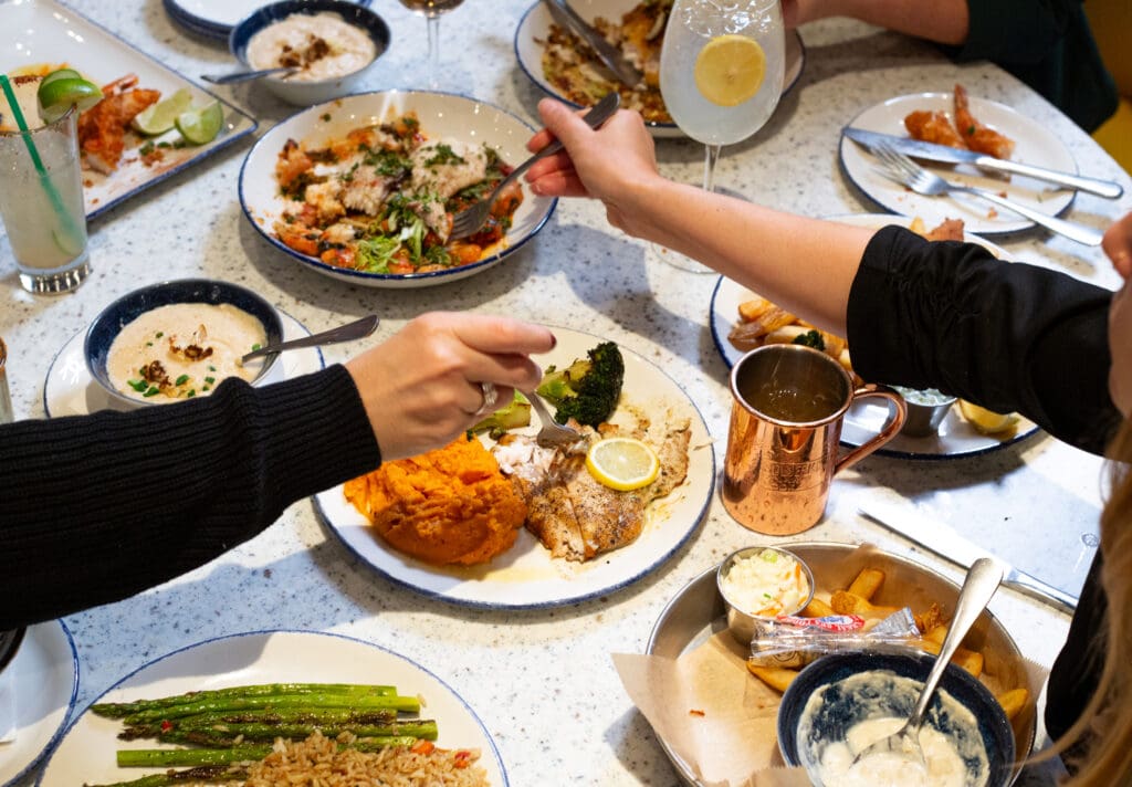 A group of people dining, with various dishes spread on the table, including fish with mashed sweet potatoes and broccoli, asparagus with rice, a salad, and a bread basket. One person is handing a drink with a lemon slice, while others are reaching for food.