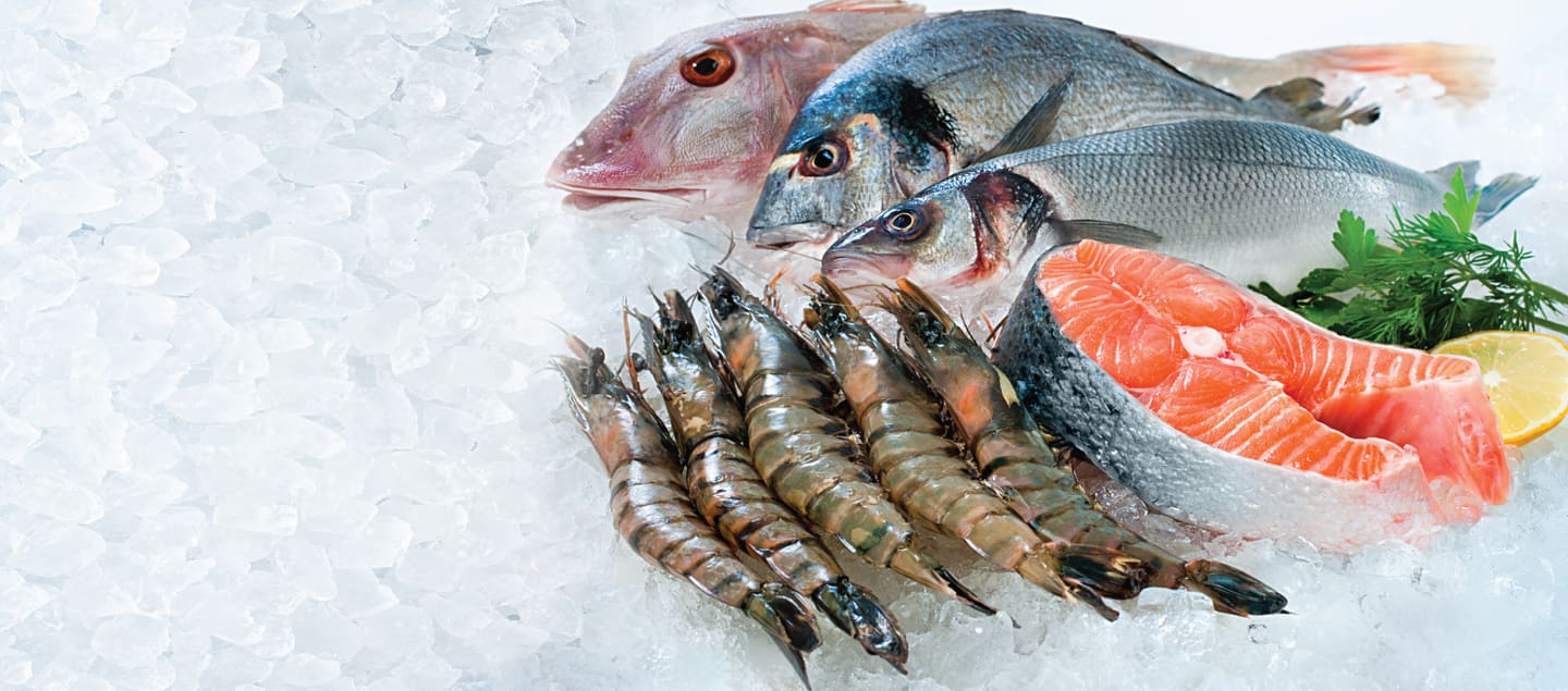 A variety of fresh seafood is displayed on ice. The selection includes several whole fish, shrimp, and salmon steaks. The seafood is accompanied by sprigs of fresh herbs and a lemon wedge.