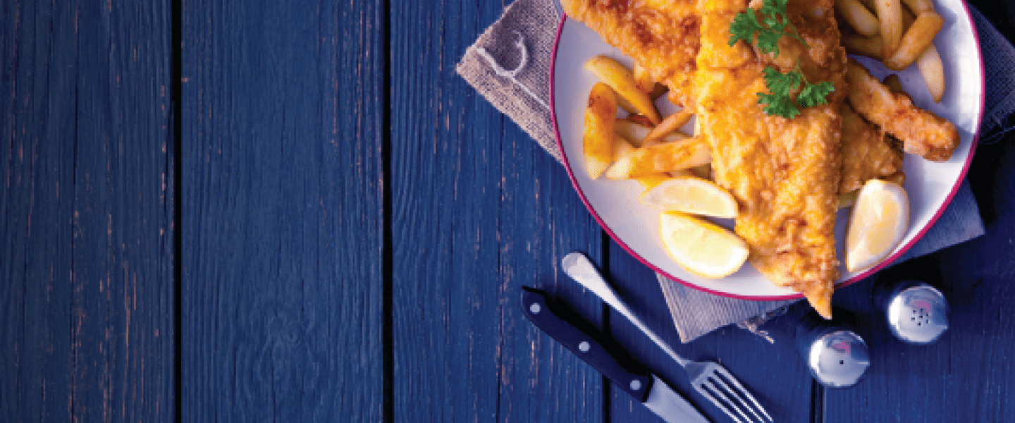 A plate of fish and chips garnished with lemon wedges and parsley sits on a dark blue wooden table. Beside the plate are a fork, a knife, and a spoon, along with salt and pepper shakers. The plate is placed on a textured beige cloth.