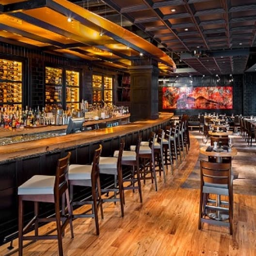 A modern restaurant interior featuring a long wooden bar lined with high chairs. To the left, shelves displaying bottles are illuminated. The dining area on the right has tables and chairs, and a vibrant, fiery-themed mural on the back wall adds ambiance.