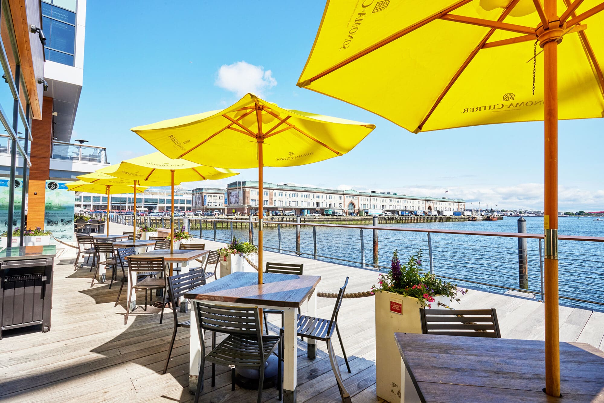 Outdoor waterfront dining area with wooden tables, metal chairs, and yellow umbrellas. The deck overlooks a calm body of water with buildings in the background. The sky is clear and sunny, creating a bright and welcoming atmosphere.