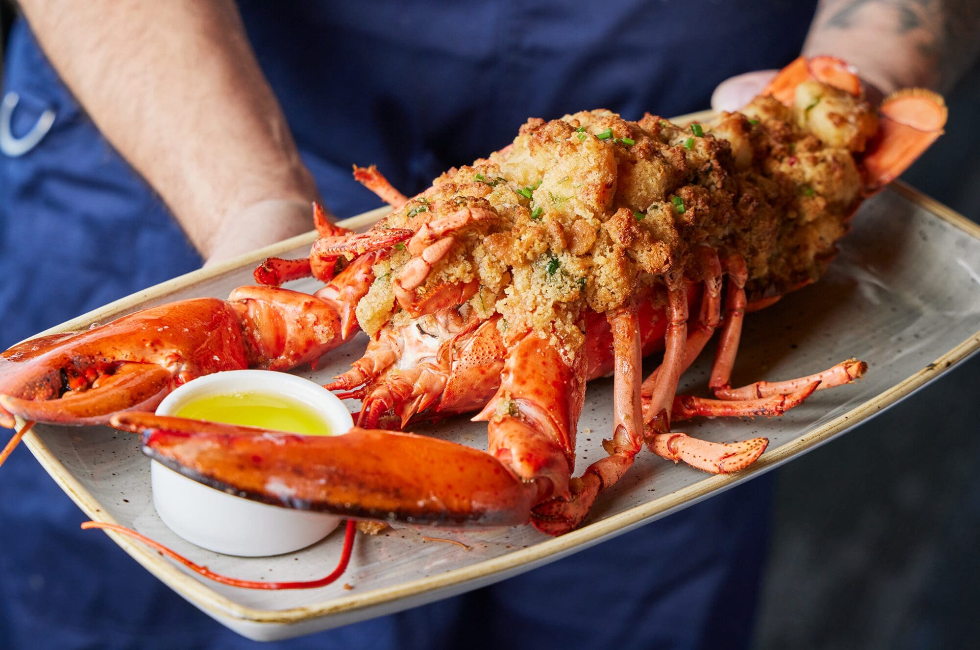 Close-up of a person holding a large platter showcasing a whole baked lobster stuffed with golden breadcrumbs and garnished with herbs. A small bowl of melted butter accompanies the dish, and the person's arms, in a blue shirt, partially frame the scene.