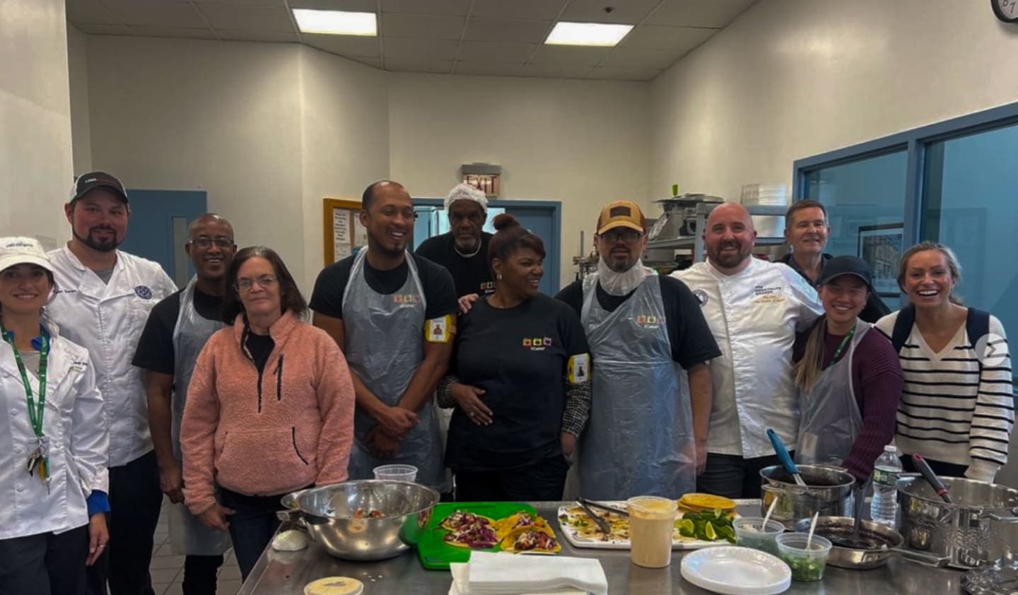 A group of twelve people, including men and women, are gathered in a commercial kitchen. Some are wearing aprons and chef's hats, indicating they are involved in cooking. The mood appears friendly and collaborative, with some people smiling at the camera.