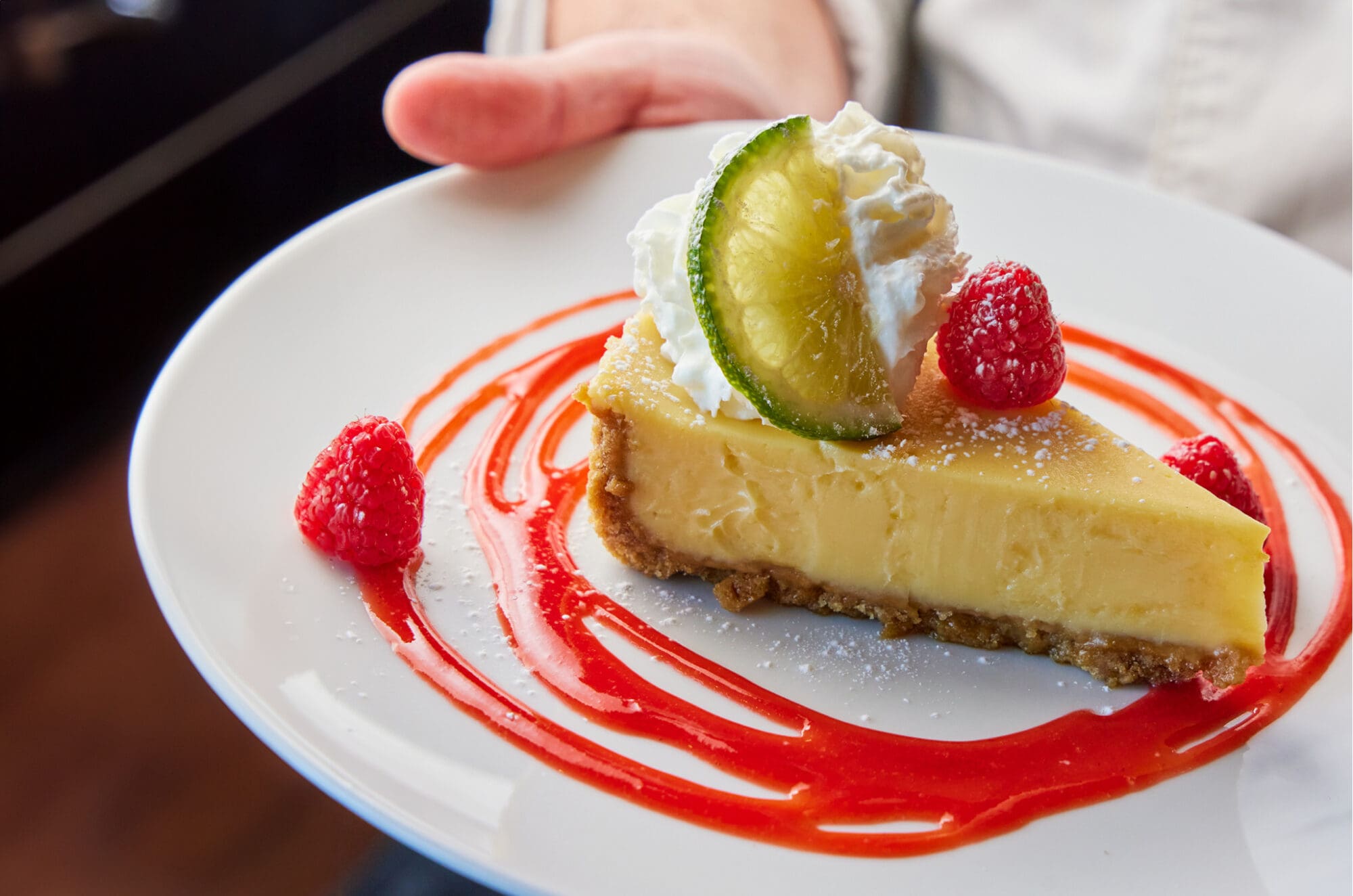 A hand holding a white plate with a slice of key lime pie garnished with a slice of lime, whipped cream, and raspberries. The pie sits on a swirl of raspberry sauce, and the crust appears crumbly and golden brown.