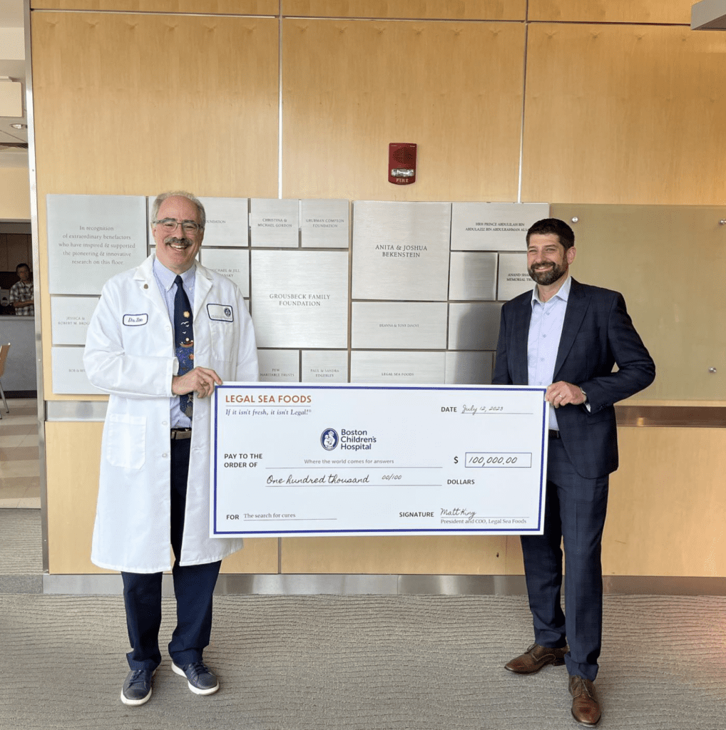 Two men are standing indoors holding a large ceremonial check. The man on the left is wearing a white lab coat, and the man on the right is dressed in a dark suit. The check is for $100,000 and is payable to 