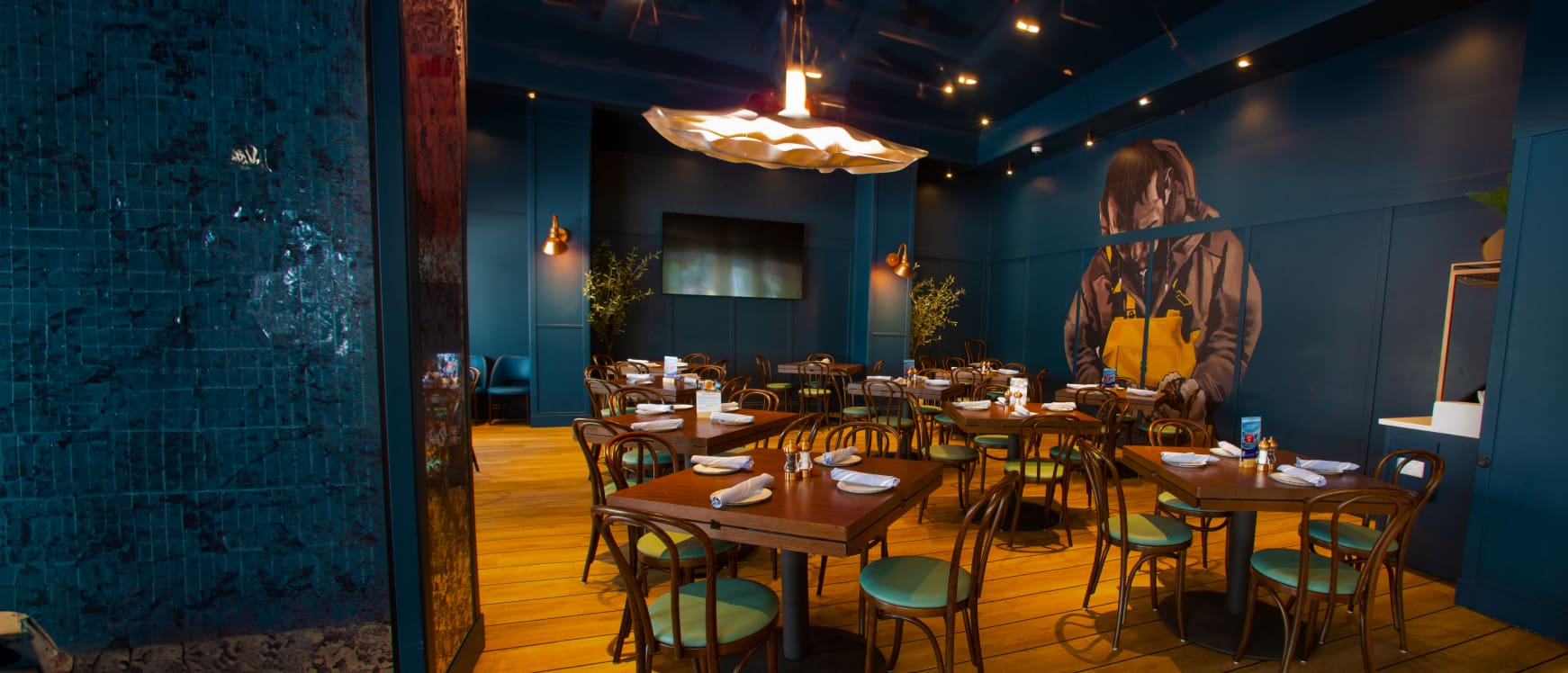 A restaurant interior with teal walls and wooden flooring. Wooden tables and chairs are neatly arranged, each table set with white napkins. A large mural of a person on the right wall and a unique ceiling light fixture add to the modern ambiance.