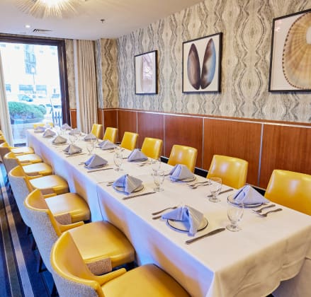 A long dining table set for an event in a private room with yellow chairs, a white tablecloth, neatly folded napkins, and glassware. The room features patterned wallpaper and framed artwork, and there is natural light coming through the large window.