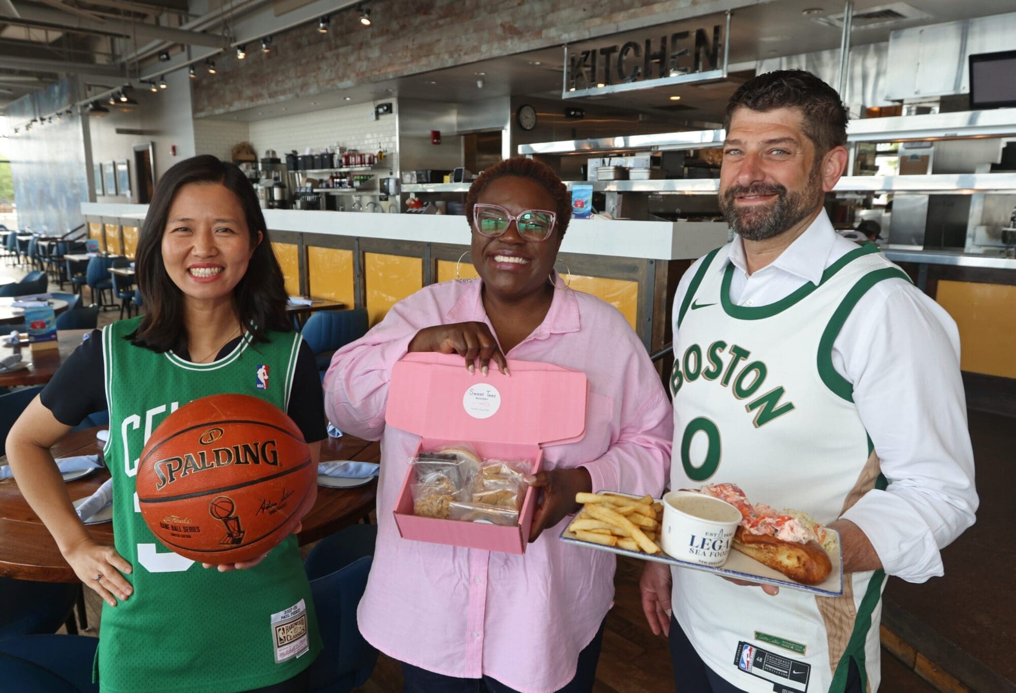 Three people smile and hold various items inside a restaurant kitchen. The person on the left holds a basketball, the person in the middle holds a box of pastries, and the person on the right holds a plate with a sandwich and fries. All are wearing Boston-themed outfits.