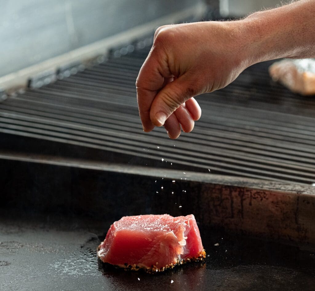 A hand is sprinkling salt over a piece of raw tuna that is placed on a hot grill. The background is slightly blurred and shows the grill's surface and grates.