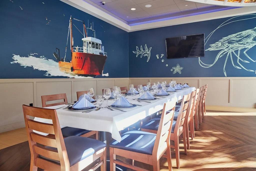 A well-lit restaurant dining area with a long table set for a meal. The walls are adorned with vivid murals of a red fishing boat on the ocean and marine life. Blue cushioned chairs surround the table, and a flat-screen TV is mounted on the wall.