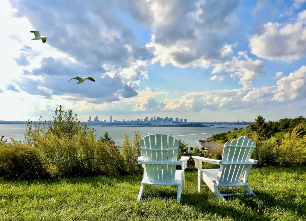 Two empty white Adirondack chairs face a scenic view of a city skyline, seen across a body of water under a partly cloudy sky. Two seagulls are flying in the air, and lush green grass and shrubs surround the chairs, creating a peaceful atmosphere.