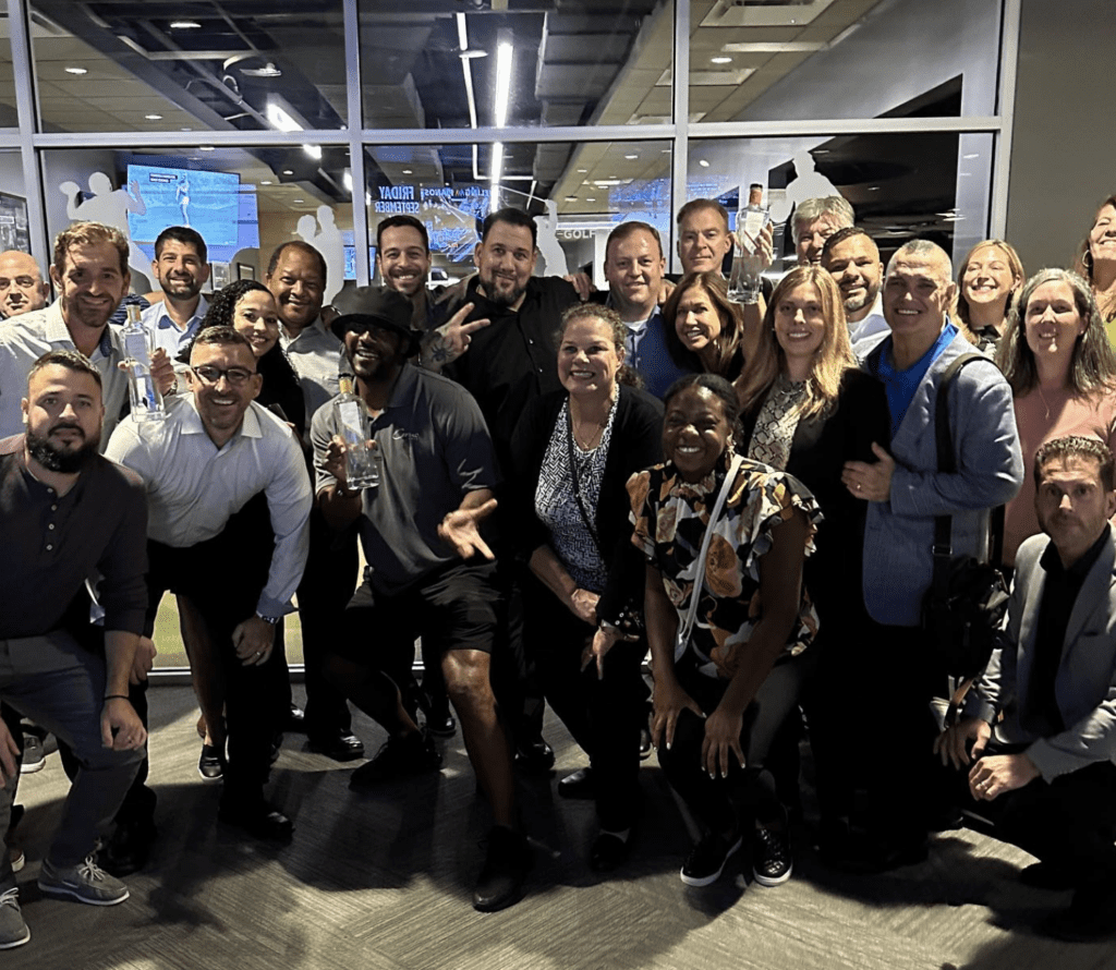 A large and diverse group of people smiling and posing indoors. Some individuals hold glass awards. The setting appears to be a modern office or conference room with glass walls, screens, and ceiling lights in the background.