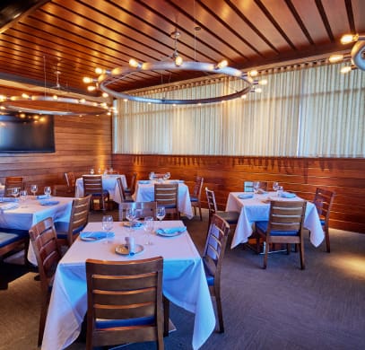 A cozy dining room features several wooden tables covered with white tablecloths and place settings. Wooden chairs surround the tables, and circular modern light fixtures hang from the wooden-paneled ceiling. The room is softly lit, creating a warm ambiance.