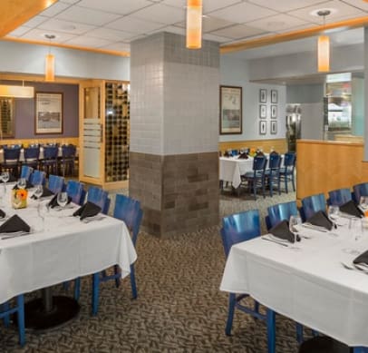 A restaurant dining area with blue chairs and white tablecloths. Tables are set with black napkins, glassware, and cutlery. The space has light gray walls, pendant lights, and framed pictures. The floor is carpeted with a light pattern, and a column divides the room.