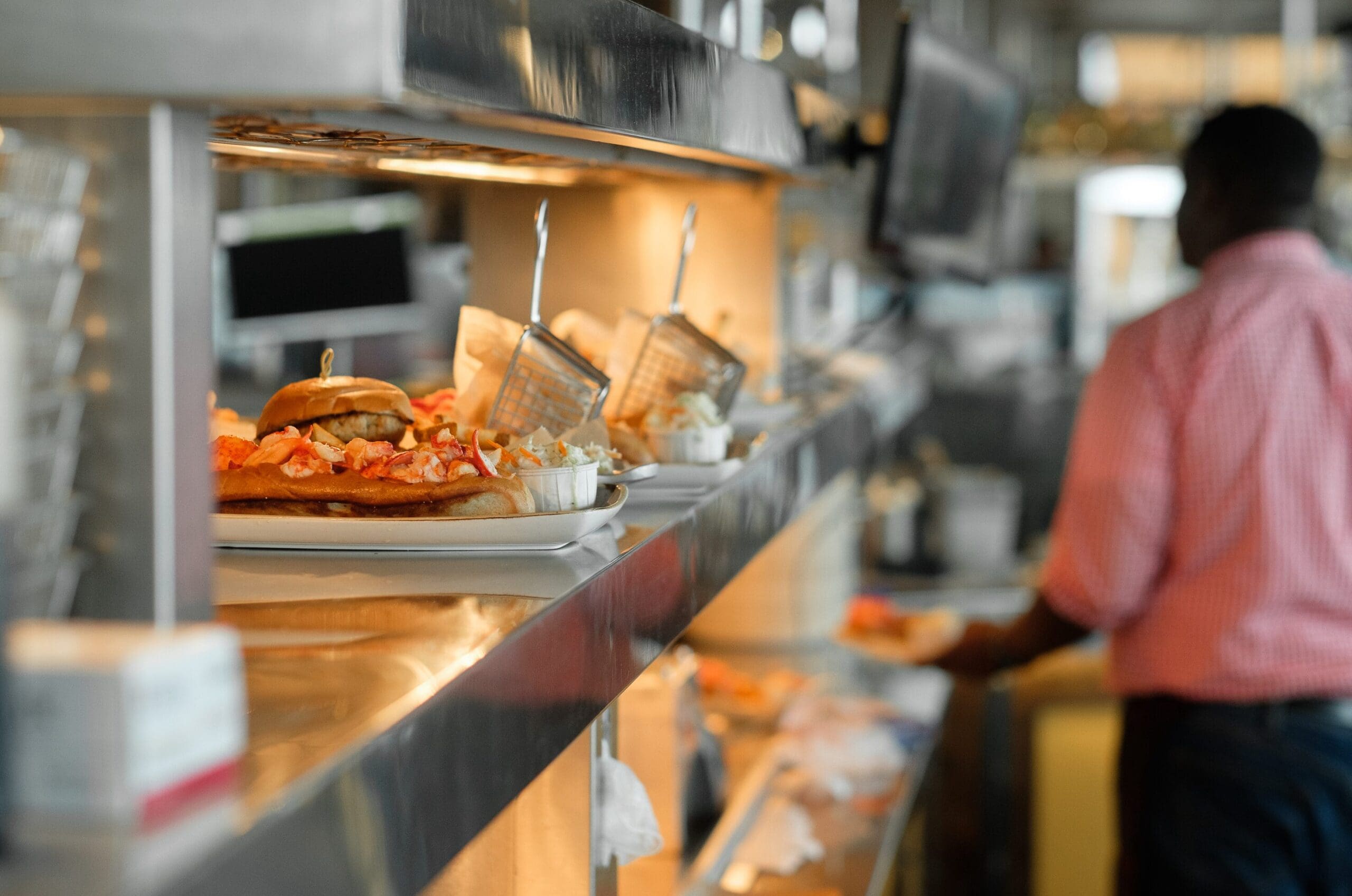 Plates of food on a kitchen pass, including a burger and fries, in a bustling restaurant. A person in a red checkered shirt is blurred in the background. The atmosphere appears busy with food preparation and service.