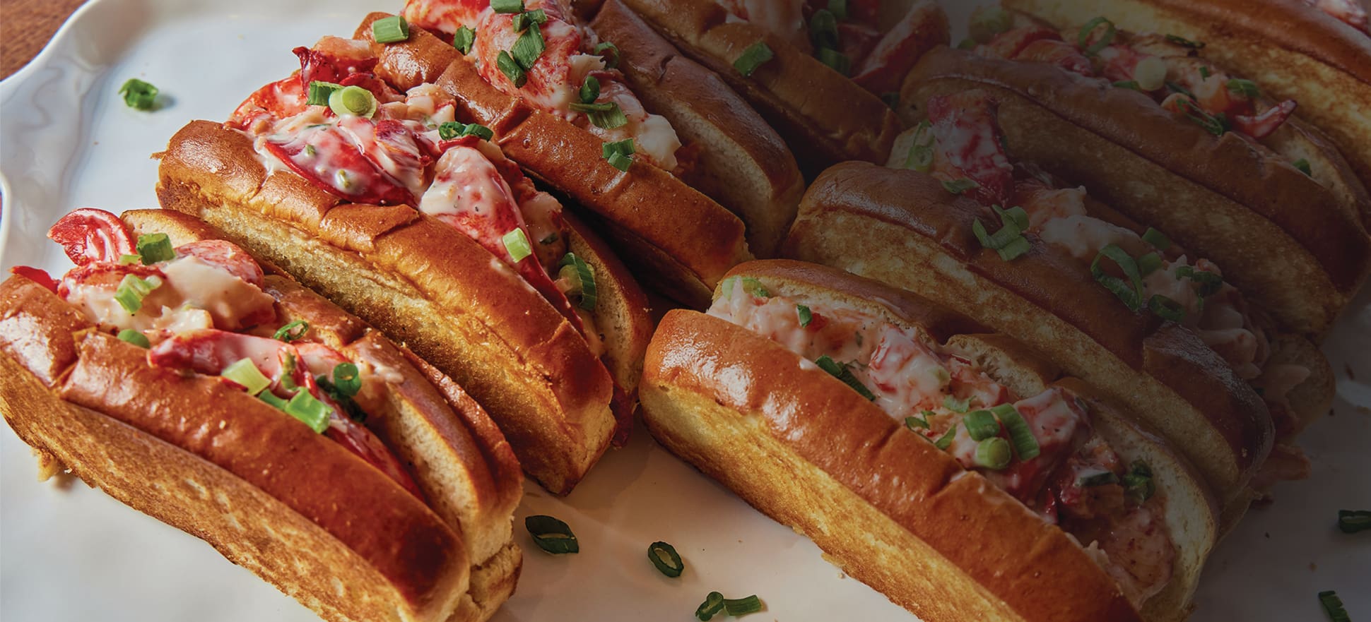 A platter of toasted lobster rolls topped with creamy lobster meat, garnished with chopped green onions. The rolls are arranged neatly on a white plate, showcasing their golden-brown sides and savory filling.