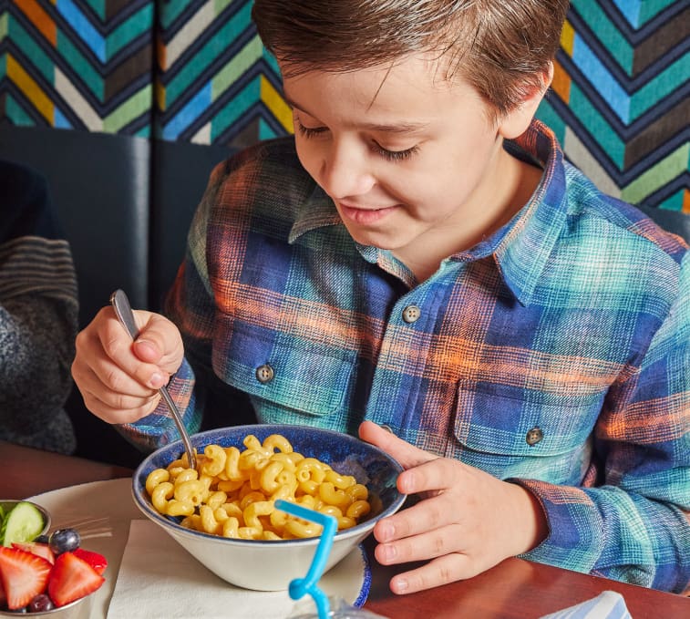 A young boy wearing a blue and green plaid shirt is sitting at a table, eating a bowl of macaroni and cheese with a fork. A plate of fresh fruit is visible on the table, and a colorful, geometric-patterned wall is in the background.