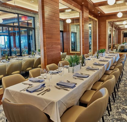 Elegant dining room with a long table set for a meal. The table is covered with a white tablecloth and adorned with wine bottles, green plants, and neatly set plates, glasses, and napkins. The room features mirrored walls, wood paneling, and modern lighting.