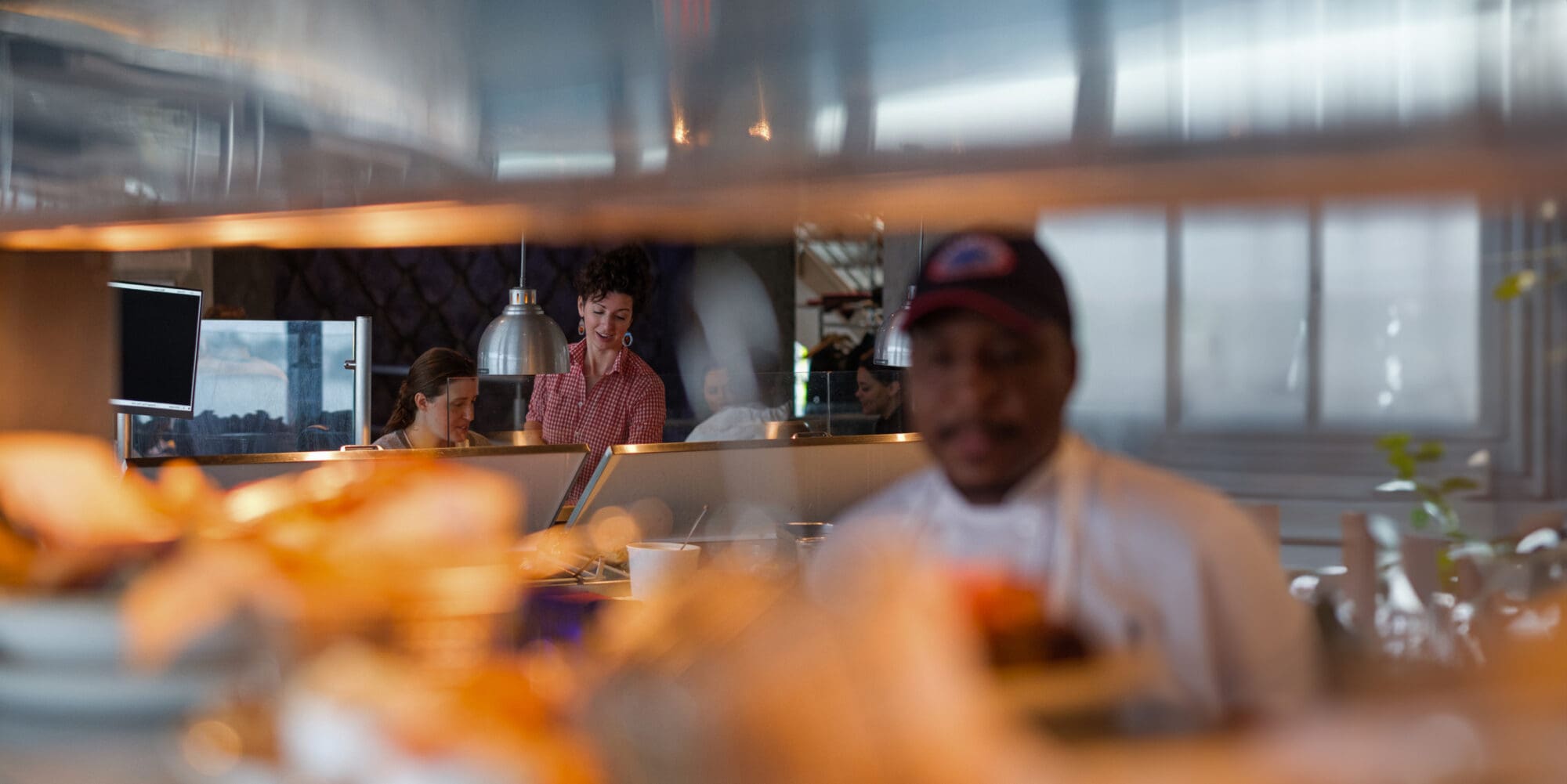 A busy restaurant kitchen with blurred food in the foreground. In the background, a chef in a white uniform and dark cap works while two people behind the kitchen counter attend to tasks. The scene has a warm, bustling atmosphere.