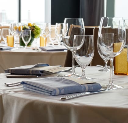 Elegant table settings with neatly arranged wine glasses, water glasses, silverware, and folded blue striped napkins on white tablecloths. Blurred background shows additional tables with similar arrangements and a window view of a city skyline.