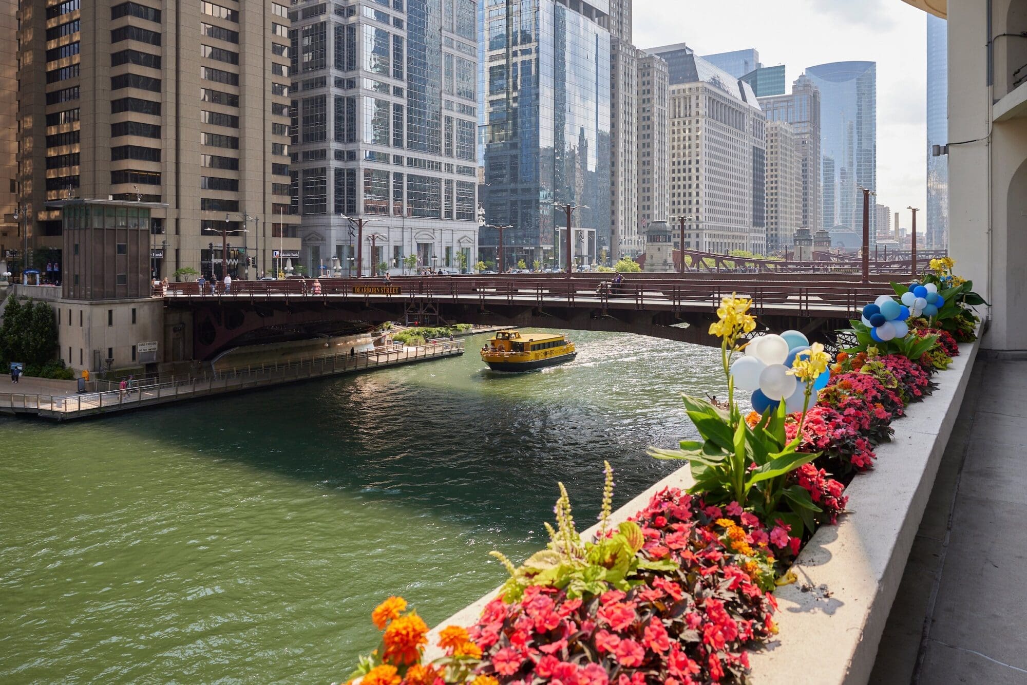 A bright yellow boat navigates a green river flanked by a bustling cityscape with tall buildings. A bridge spans the river, and in the foreground, colorful flowers and blue and white balloons adorn a parapet. The sun shines brightly on this urban scene.