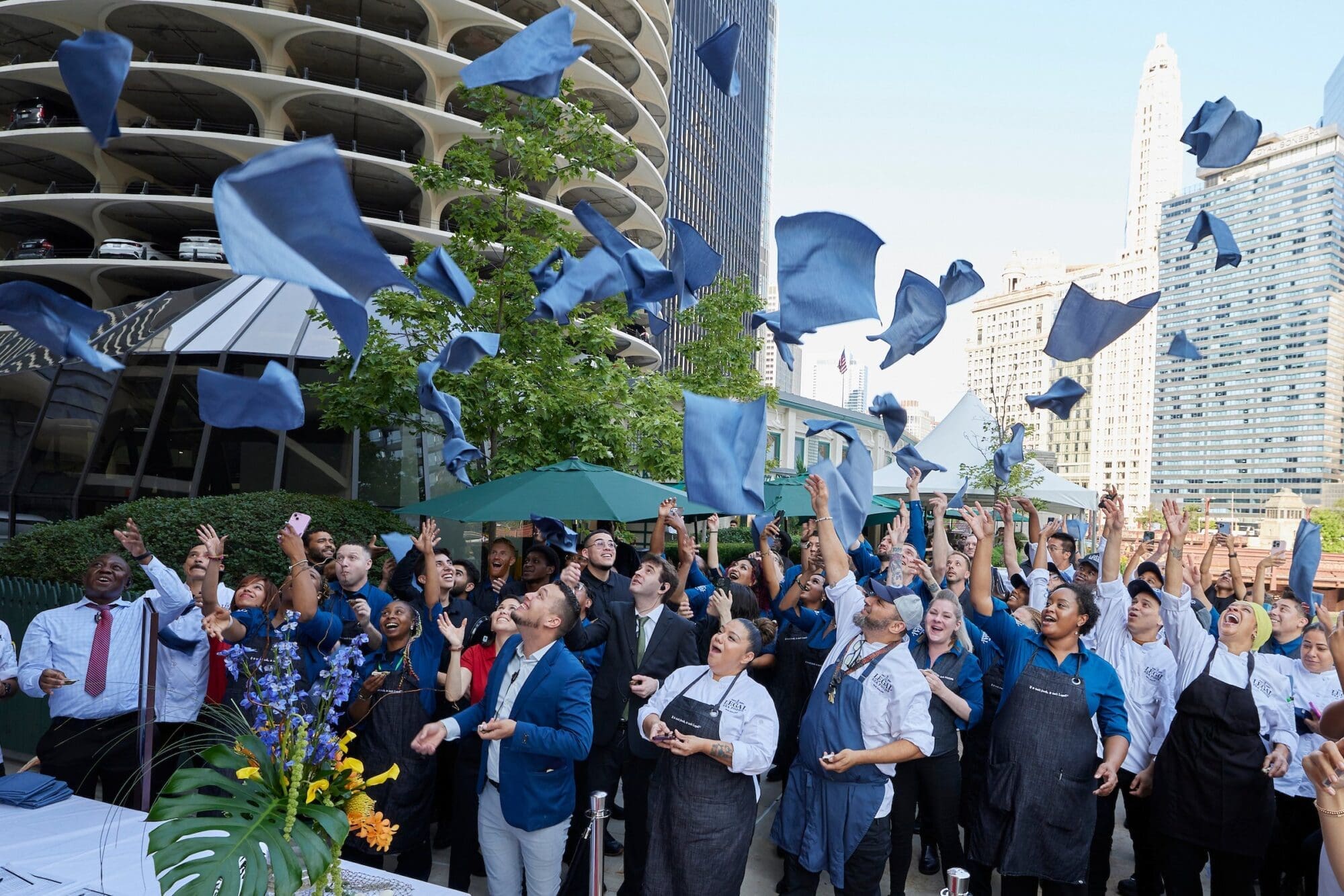 A large group of people, including individuals in chef uniforms and others in business attire, cheer and throw blue aprons into the air in an outdoor setting, surrounded by tall buildings and greenery. The mood is celebratory and festive.