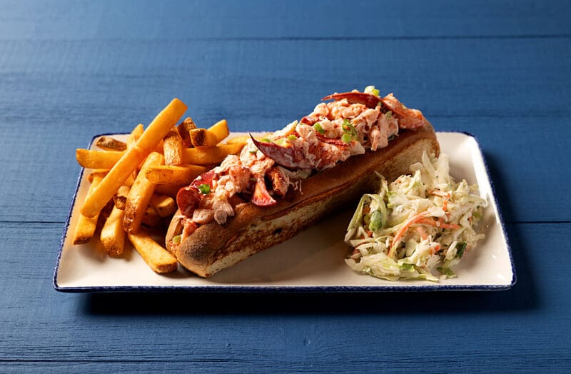 A rectangular plate on a blue surface holds a lobster roll topped with chunks of lobster meat and green garnish in a toasted bun. On one side of the roll are golden French fries, and on the other side is a serving of coleslaw.