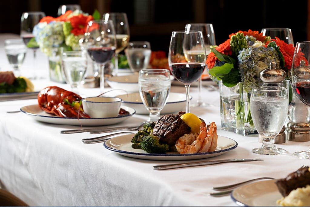 A beautifully set dining table adorned with floral centerpieces and place settings. Dishes include surf and turf with shrimp, steak, vegetables, and lobster. Glasses of red wine and water accompany the meals, creating an elegant dining atmosphere.