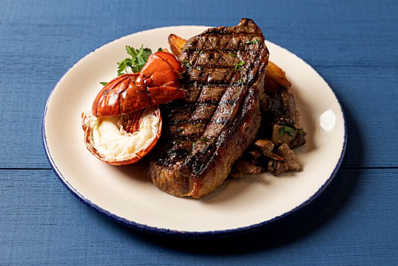 A grilled steak and half a lobster tail are served on a white plate with a blue rim. They are accompanied by roasted vegetables and garnished with fresh parsley. The background is a blue wooden surface.