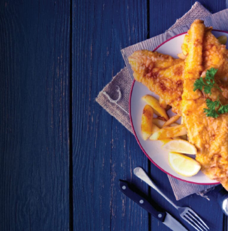 A white plate with a red rim holds a serving of golden-brown fried fish fillets, garnished with parsley and accompanied by lemon wedges and fries. The plate rests on a piece of burlap on a blue wooden table. A knife and fork are placed beside the plate.